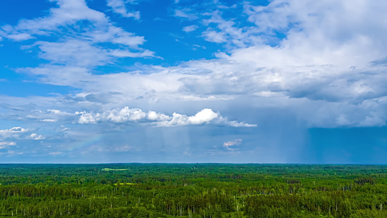 A colorful rainbow shines over the green, European, rural landscape during light rain - high altitude aerial time lapse and flowing cloudscape