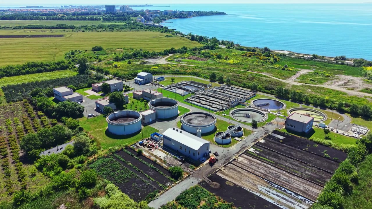 Water treatment facility located near the coast with tanks and buildings surrounded by green fields and the sea in the background on a bright day