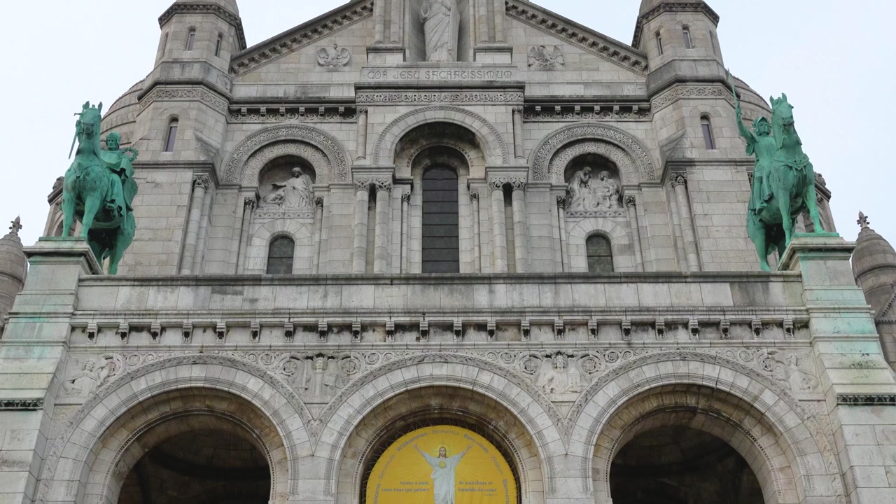 Frontal symmetrical view of the Sacré-Cœur Basilica in Montmartre, Paris. The facade features arches, sculptures, domes, and the iconic green equestrian statues on both sides under a cloudy sky