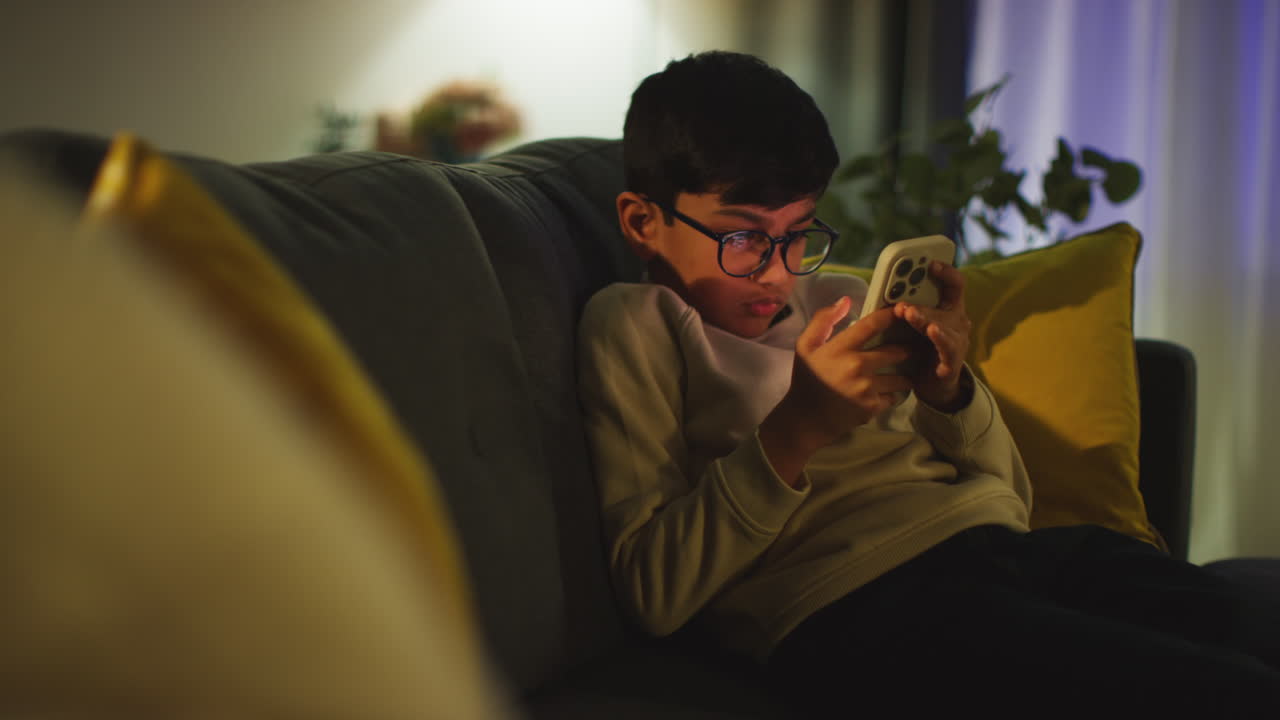 Young Boy Sitting On Sofa In Lounge At Home Playing Game On Mobile Phone At Night 2