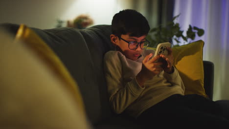 Young-Boy-Sitting-On-Sofa-In-Lounge-At-Home-Playing-Game-On-Mobile-Phone-At-Night-2