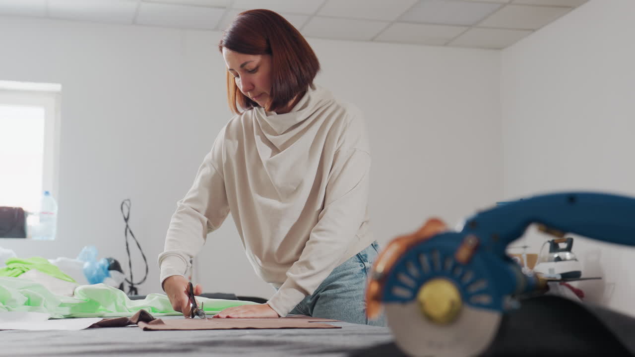 Seamstress working in sewing workshop, holding fabric down with one hand while cutting out design pattern with scissors, surrounded by sewing materials, machine tools, and fabric on wide table