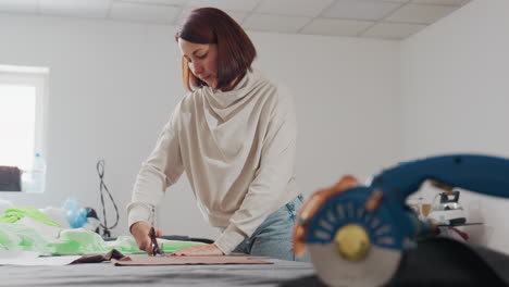 seamstress working in sewing workshop, holding fabric down with one hand while cutting out design pattern with scissors, surrounded by sewing materials, machine tools, and fabric on wide table