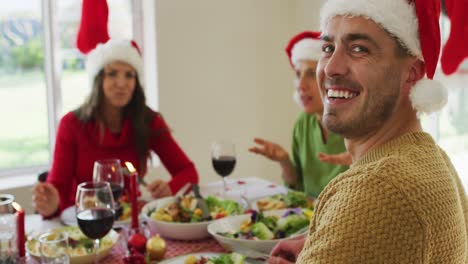 Feliz-Hombre-Caucásico-Con-Sombrero-De-Papá-Noel-Mirando-La-Cámara-Durante-La-Comida-De-Navidad
