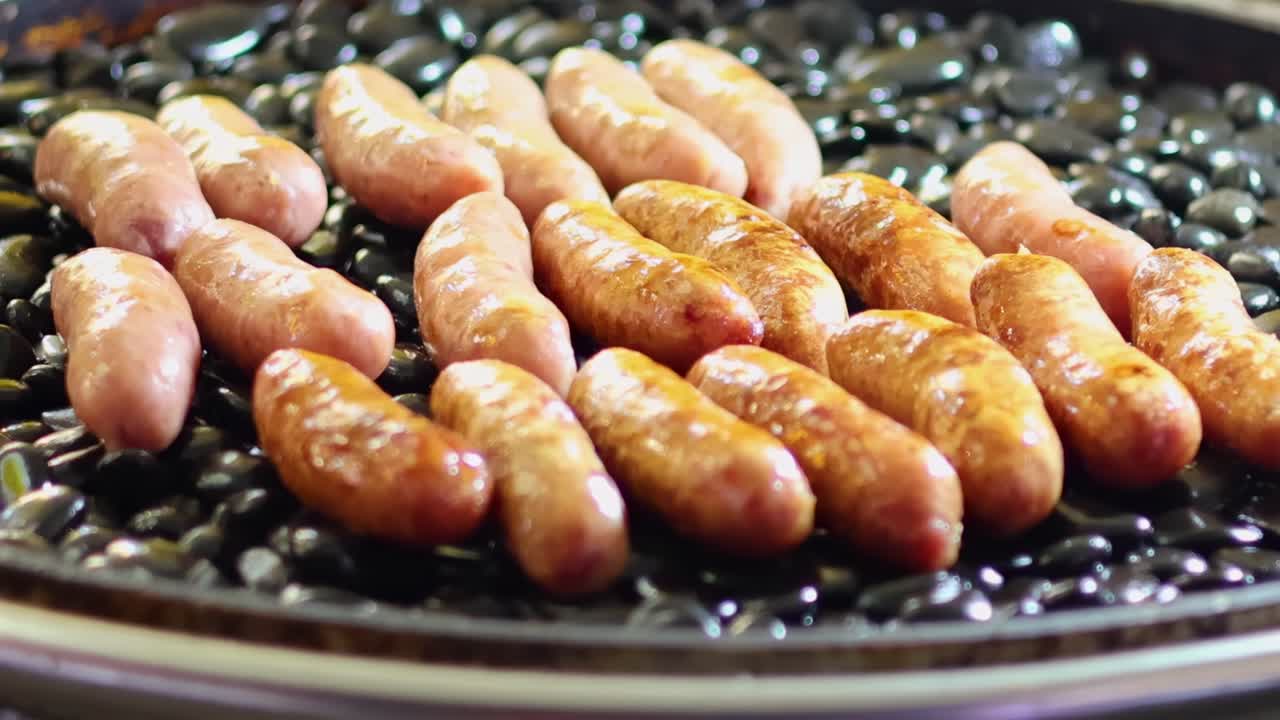 Close-up of sausages cooking on a rotating grill over black stones, showcasing a savory grilling process.