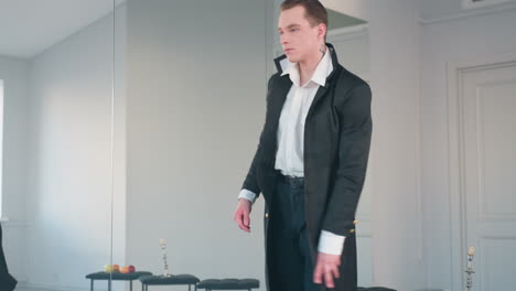 man in formal outfit performs expressive dance in bright room, bowing forward with intense movement near closed door, large mirror reflecting background with apples and candle placed on black stools