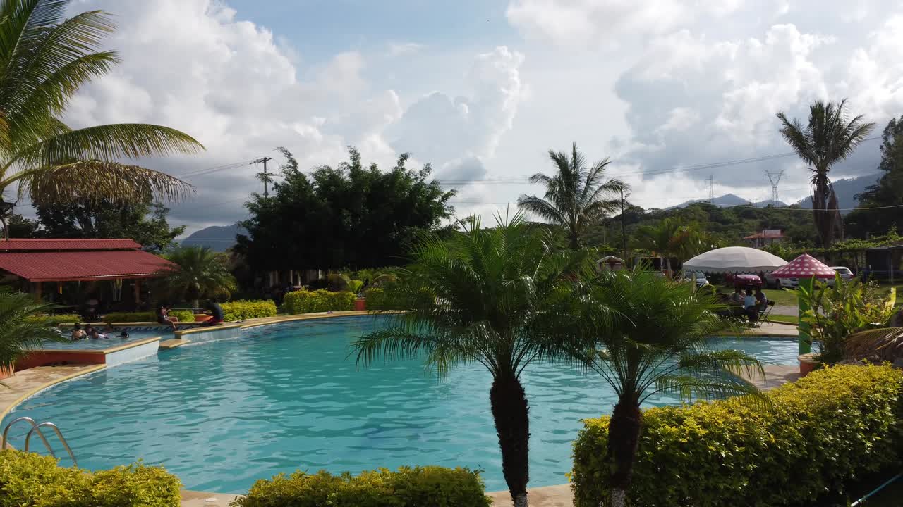 Resort pool area surrounded by palm trees, trimmed bushes, and distant mountains under bright daylight with blue sky and scattered clouds in tranquil countryside setting in a French province