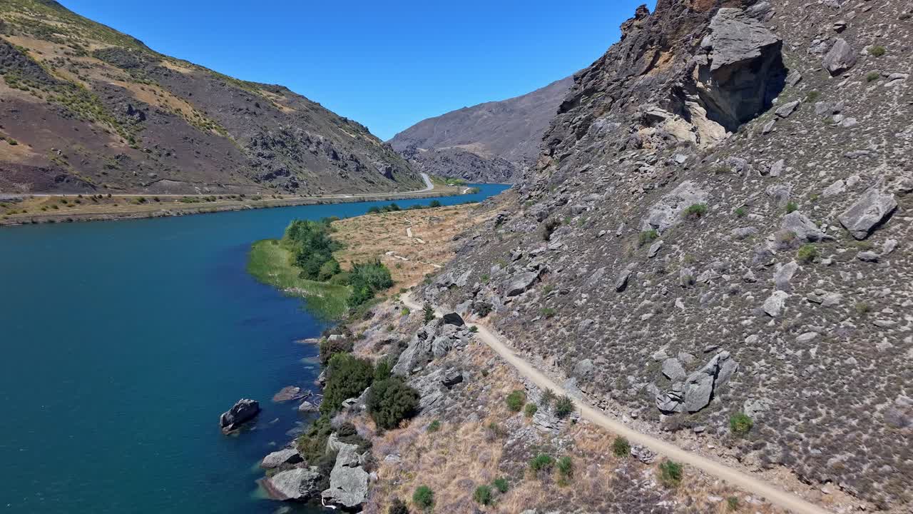 On a bright sunny day, a drone approaches the Dunstan Trail in Central Otago, showcasing rugged rocky hills, the winding path, and part of Lake Dunstan