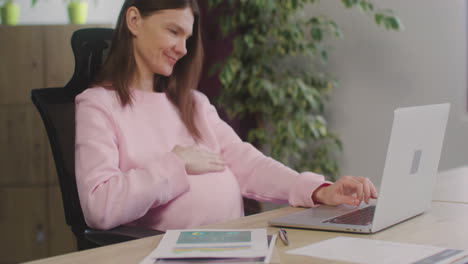 happy pregnant woman caressing her belly and drinking water while working in the office