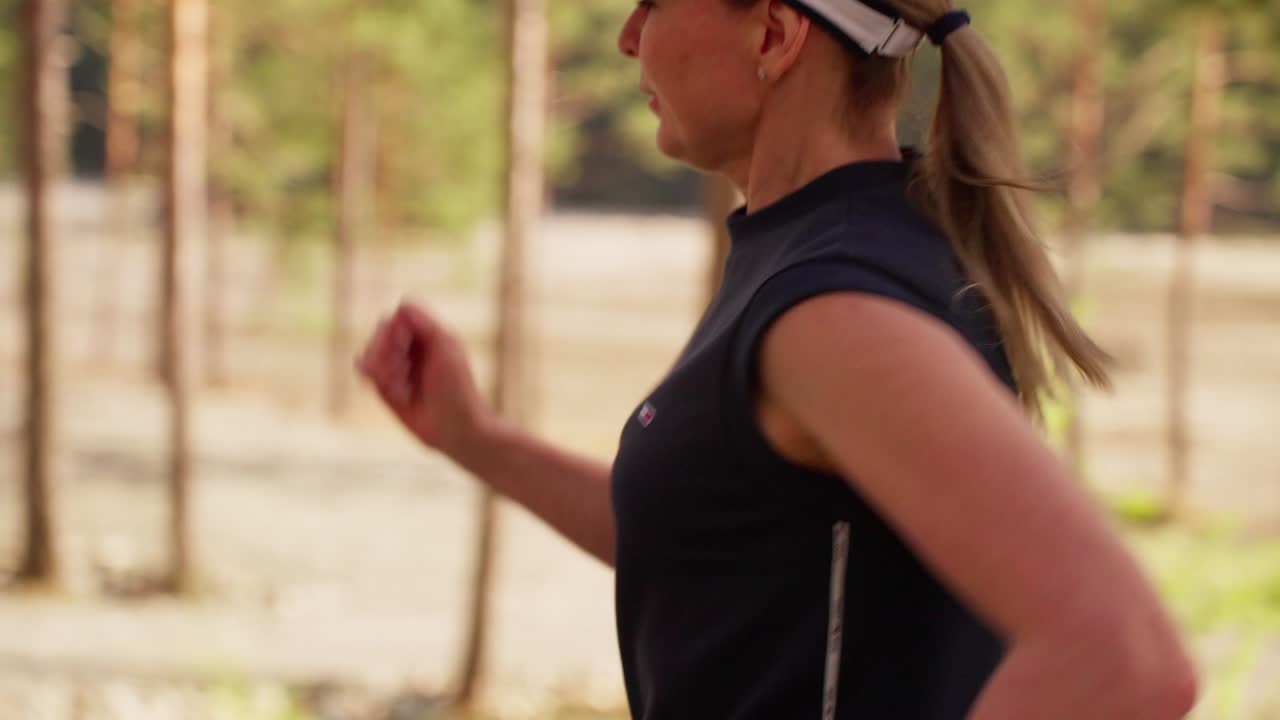Una donna cammina attraverso una foresta, la sua coda di cavallo ondeggia ad ogni passo. La luce del sole filtra attraverso alberi alti, proiettando ombre morbide sul suo percorso. Indossa un cappellino e un top sportivo scuro. La sua espressione è calma.