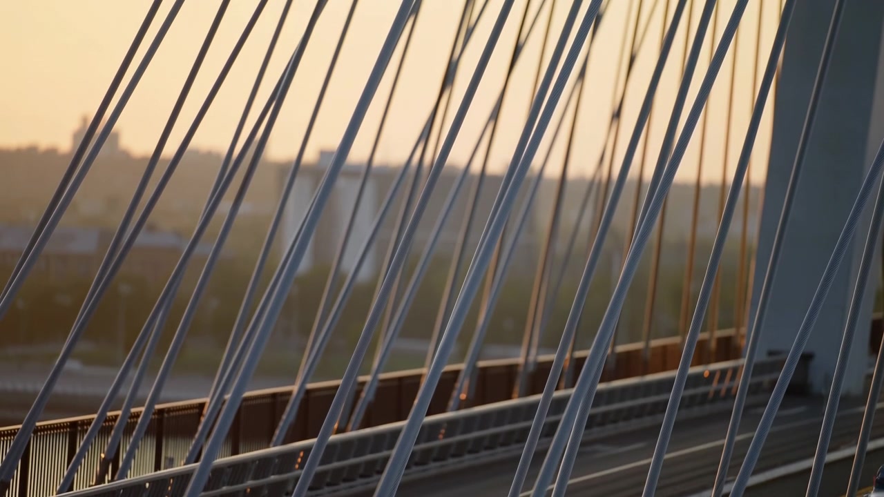 Diagonal steel suspension cables and a support pylon dominate the foreground while a roadway with lane markings, railings, and barriers runs along the lower