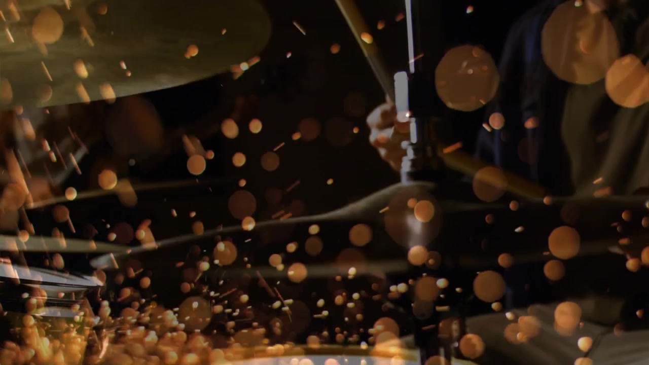 Drummer's hands entering with sticks striking snare and cymbal making music and warm particles rise