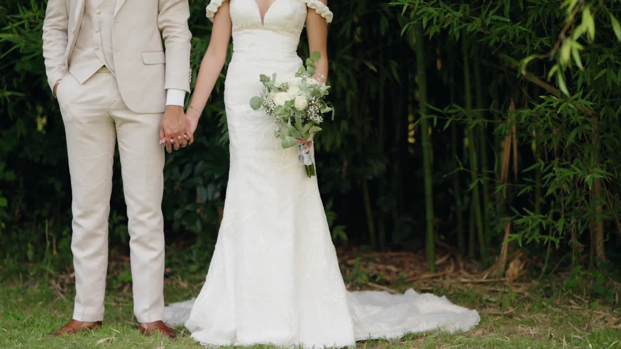 la novia en vestido de encaje blanco y el novio en traje beige sosteniendo las manos delante del bambú
