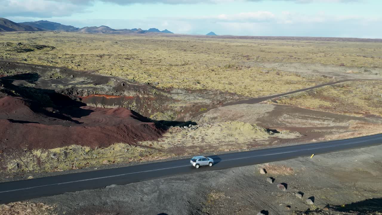 volando alto sobre islandia, viendo un vehículo navegar por un terreno de musgo y rocas volcánicas, con montañas cubiertas de nieve que se elevan contra un cielo azul profundo, despertando el deseo de viajar