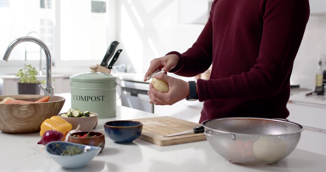 African American man opening compost bin, selecting potato, peeling it at bright kitchen island