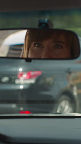 reflection of woman in rear view mirror on windshield in car. female driver puts hands on steering wheel looking to sides standing at red light closeup