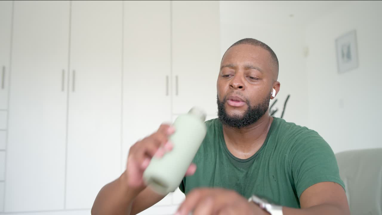 Voksen afrikansk mand gisper efter træning på gulvet, i grøn T-shirt, taler i hørepropper, mens han drikker af en flaske.