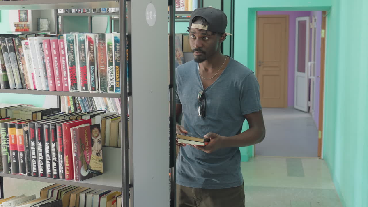 Joven vestido de forma casual de pie entre estanterías en una biblioteca, seleccionando cuidadosamente un libro de cubierta marrón de la estantería con expresión decidida, rodeado de lomos coloridos y un fondo luminoso.