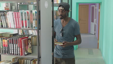 young man in casual wear standing between bookshelves in library, carefully selecting brown cover literature from shelf with intent expression, surrounded by colorful spines and bright background