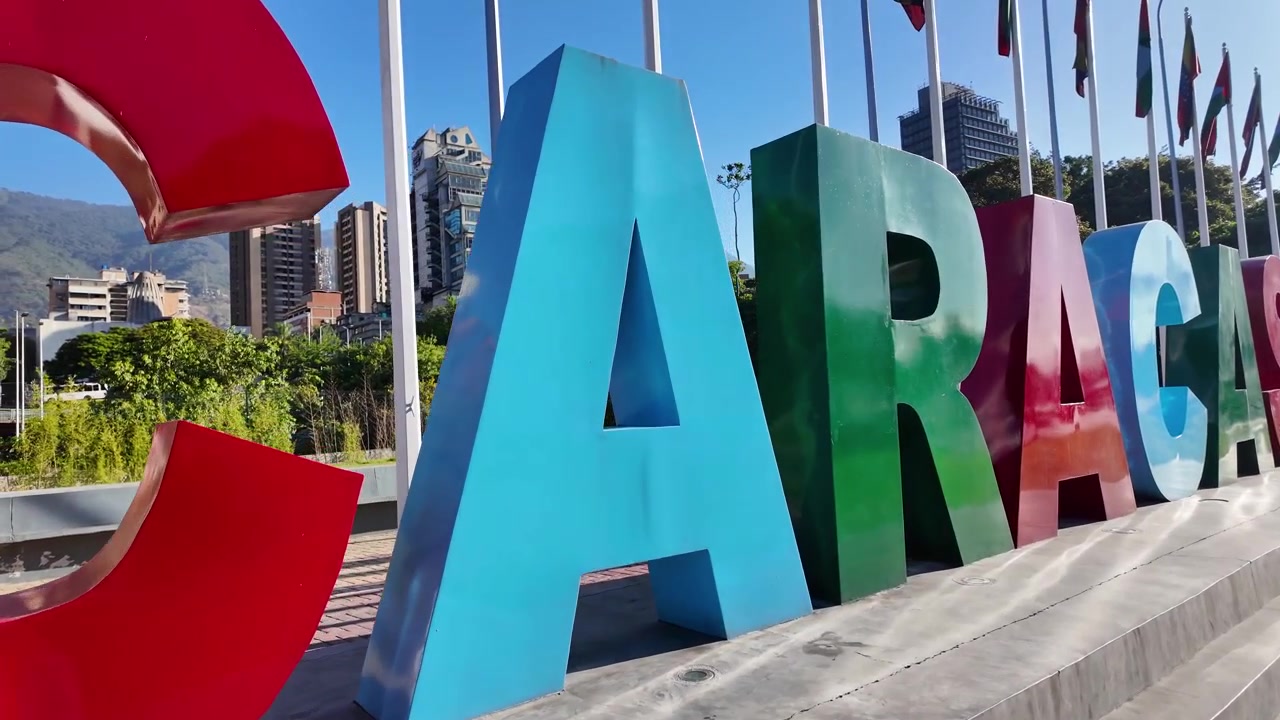 Carrellata fluida che rivela le gigantesche lettere colorate "CARACAS" a Plaza Venezuela, mostrando lo skyline urbano e l'iconica montagna Waraira Repano sotto un cielo azzurro e limpido.