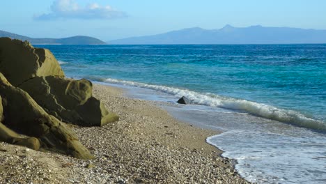 paradise beach with pebbles and rocks washed by waves of blue turquoise sea with corfu island in background