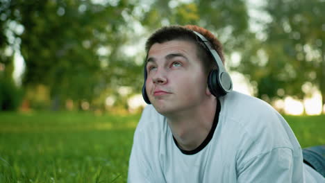 close up of artist lying on grass wearing headphones nodding to music while using phone, gazing up with a subtle smile, surrounded by lush greenery and soft bokeh effect in the serene background