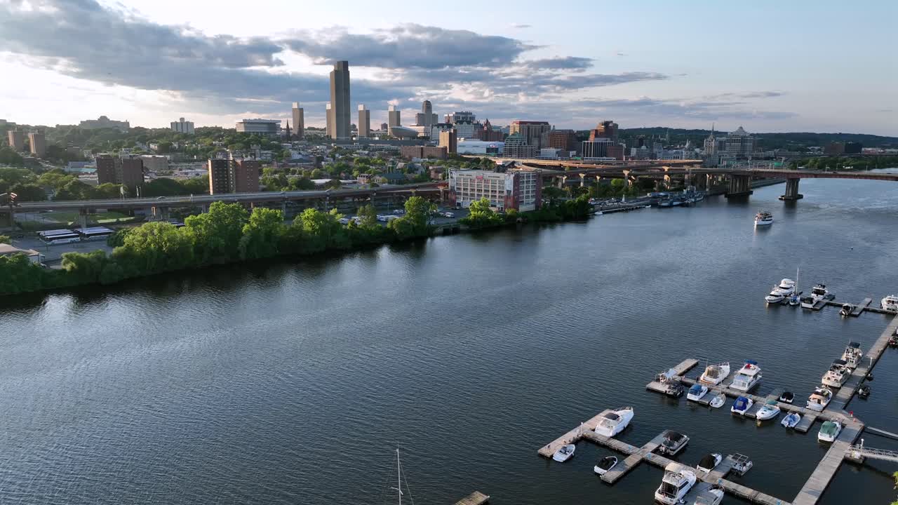 Docking yachts and boats on Hudson river at sunset. Aerial descend wide shot. Albany Yacht club and skyline of downtown in background. Traffic on coastal interstate road in New York