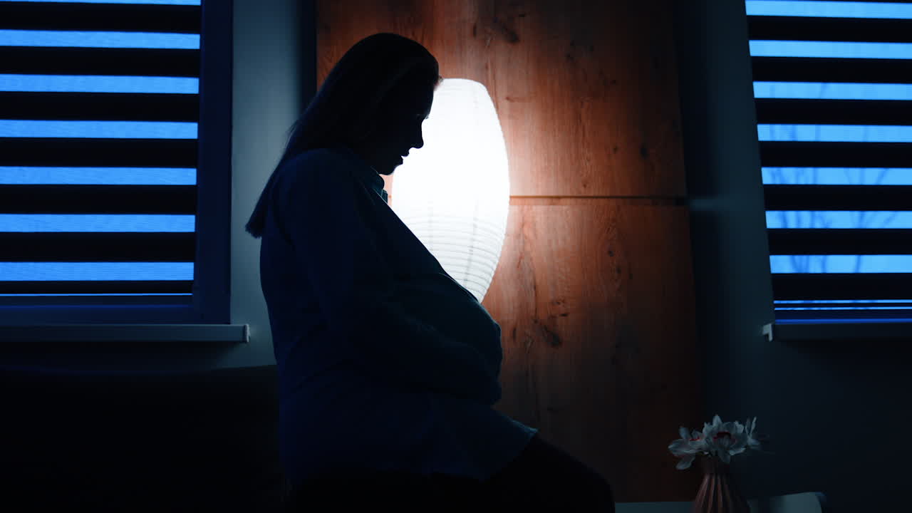 Silhouette of pregnant female sitting on the bed holding by the belly. Worried future mom in a dark room.