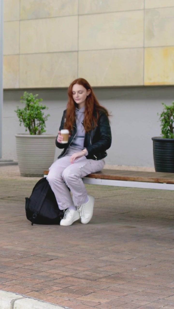 Video verticale: Donna in attesa con giacca scura che guarda l'orologio su una panchina in piazza, sorseggiando una tazza.