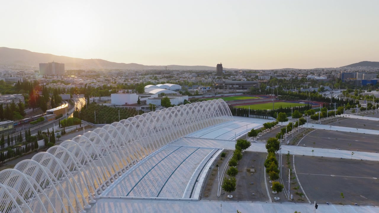 Majestic aerial footage of the Calatrava Agora arches at Athens Olympic Sports Complex at sunrise. Modern architecture glows in soft morning light above empty avenues and parking lots.