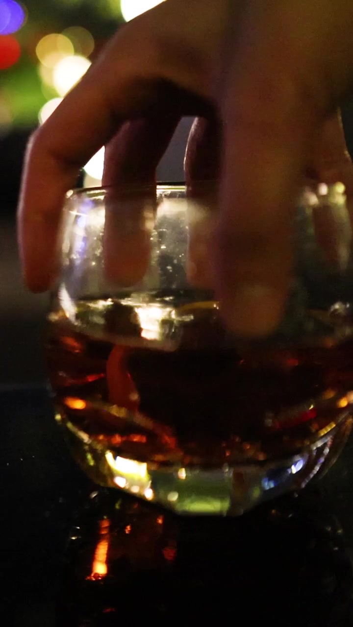 En hånd rækker ud efter og løfter et cocktailglas fyldt med mørk spiritus fra en reflekterende baroverflade, efterladende en kondensring. Varm, dæmpet belysning og bokeh-baggrund skaber en stemningsfuld nattelivsatmosfære