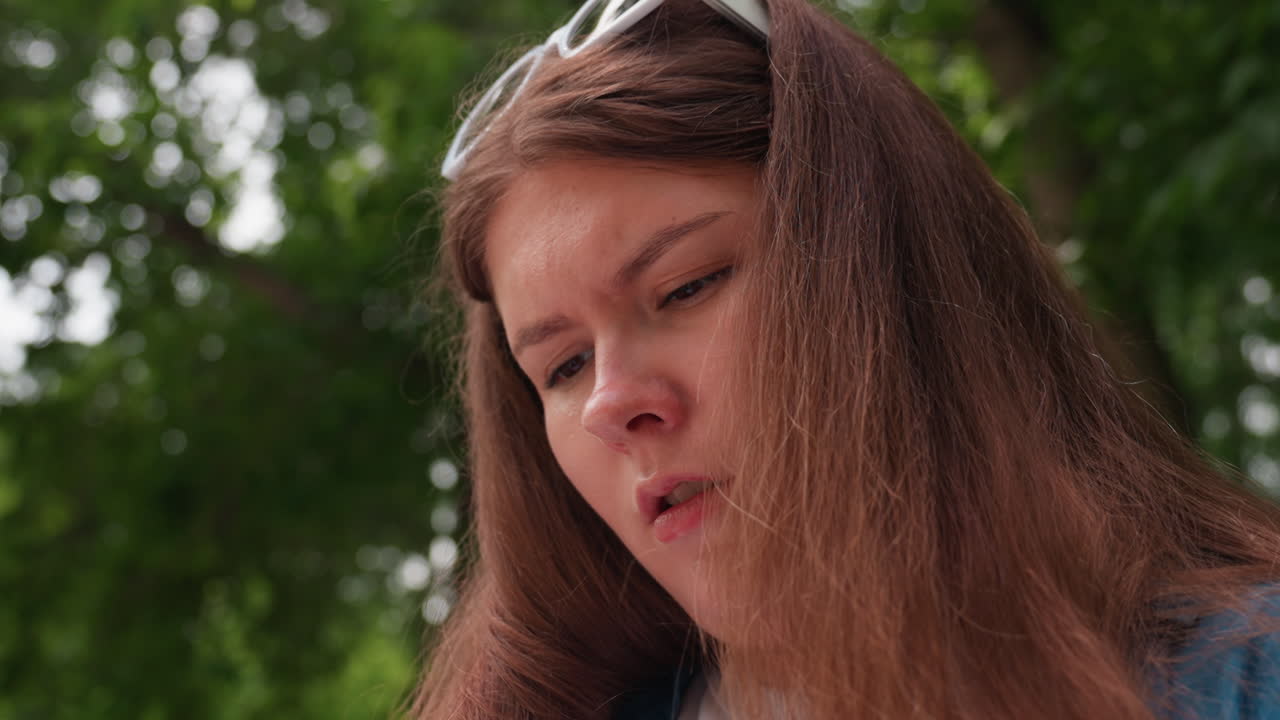 Close up focused lady with glasses on head working outdoors, concentrated face framed by brown hair, intent gaze while managing craft task in park under soft daylight