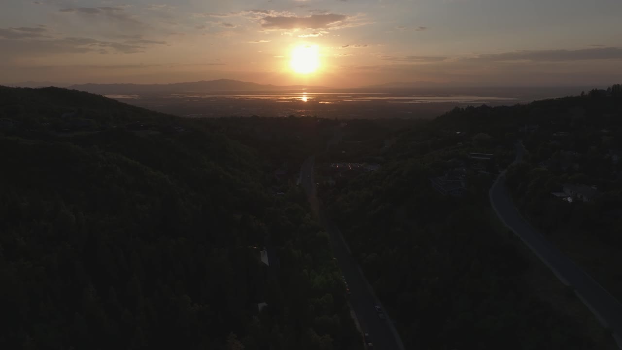 Aerial drone dolly in tilt up shot from Bountiful Canyon in Utah at golden hour with road, forest, valley homes, Wasatch Mountains, cityscape, and the Great Salt Lake at sunset