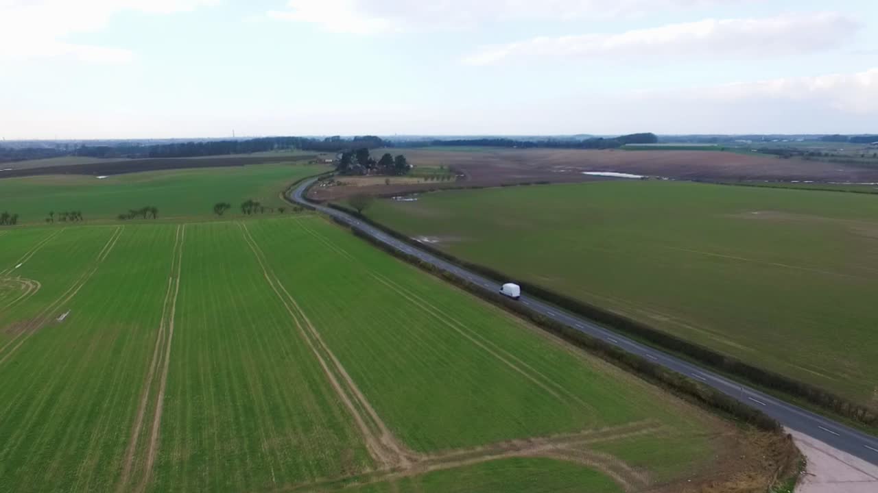 Stationary drone shot of a road as cars pass. A white van drives down the road between the fields