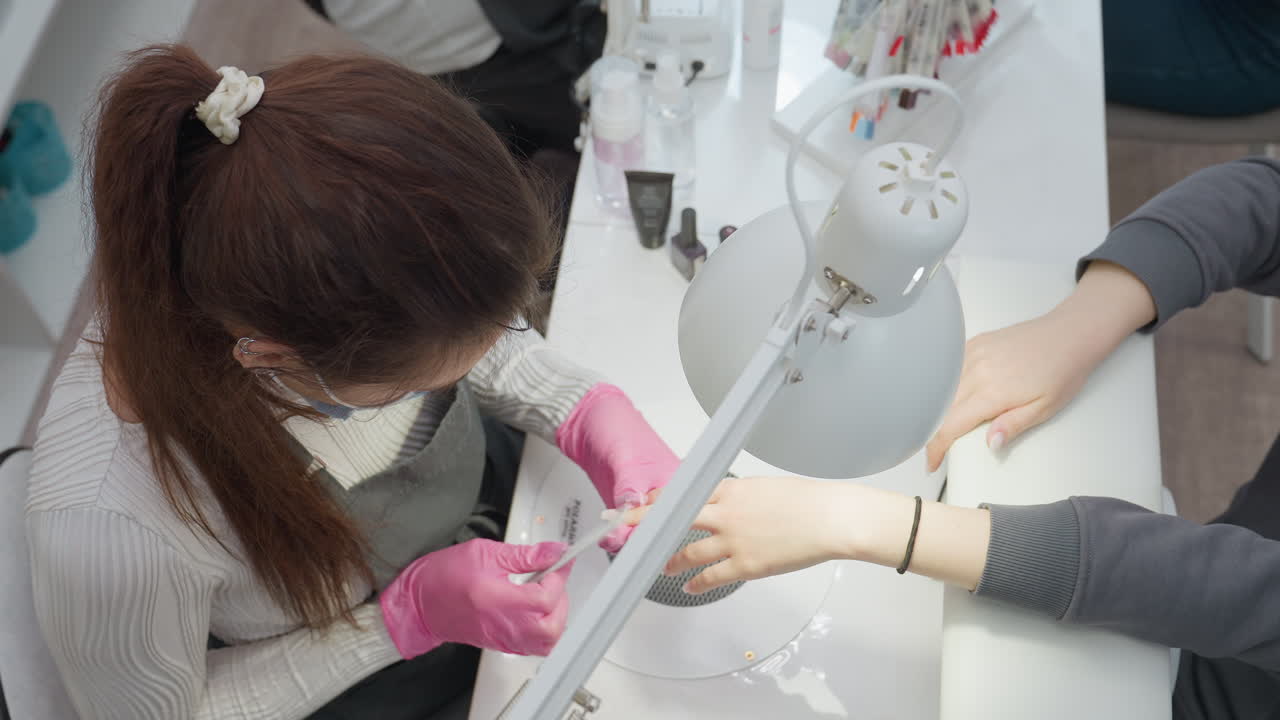 Overhead view of nail technician wearing pink gloves carefully filing client nails at well-equipped manicure station, with hand tools, nail products, and bright task lamp visible on white workspace
