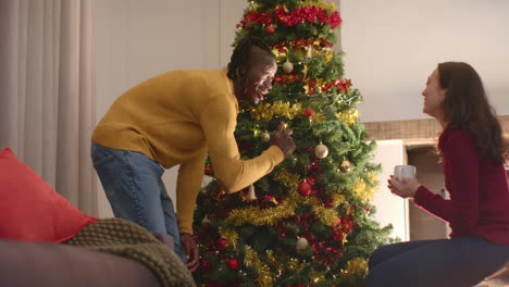 una pareja feliz y diversa tomando café y decorando el árbol de navidad en casa, en cámara lenta.