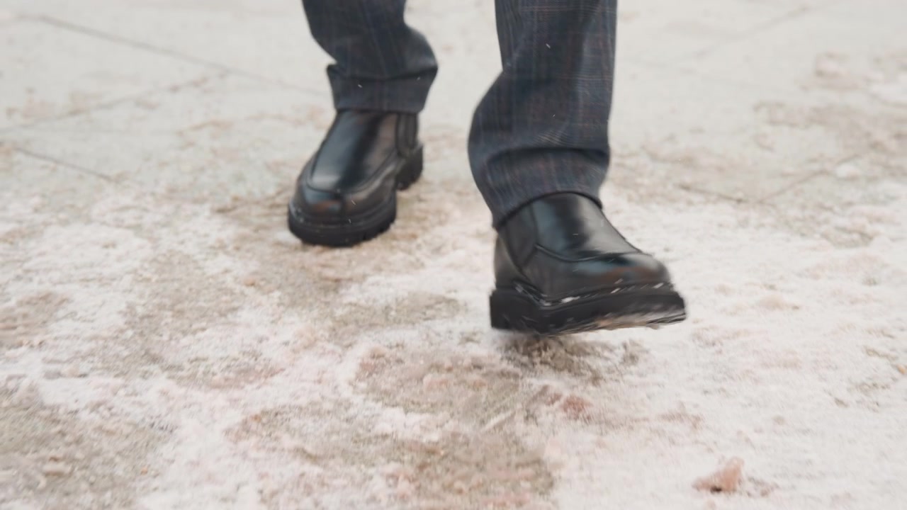 Close up of man wearing black leather shoes and dark plaid pants walking on wet concrete pavement. man feet move steadily showing soles making contact with water covered ground. Lighting
