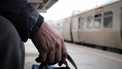 an older individual sits at a train station, clutching a brown bag with weathered hands, waiting as raindrops fall softly. the atmosphere is quiet and contemplative.
