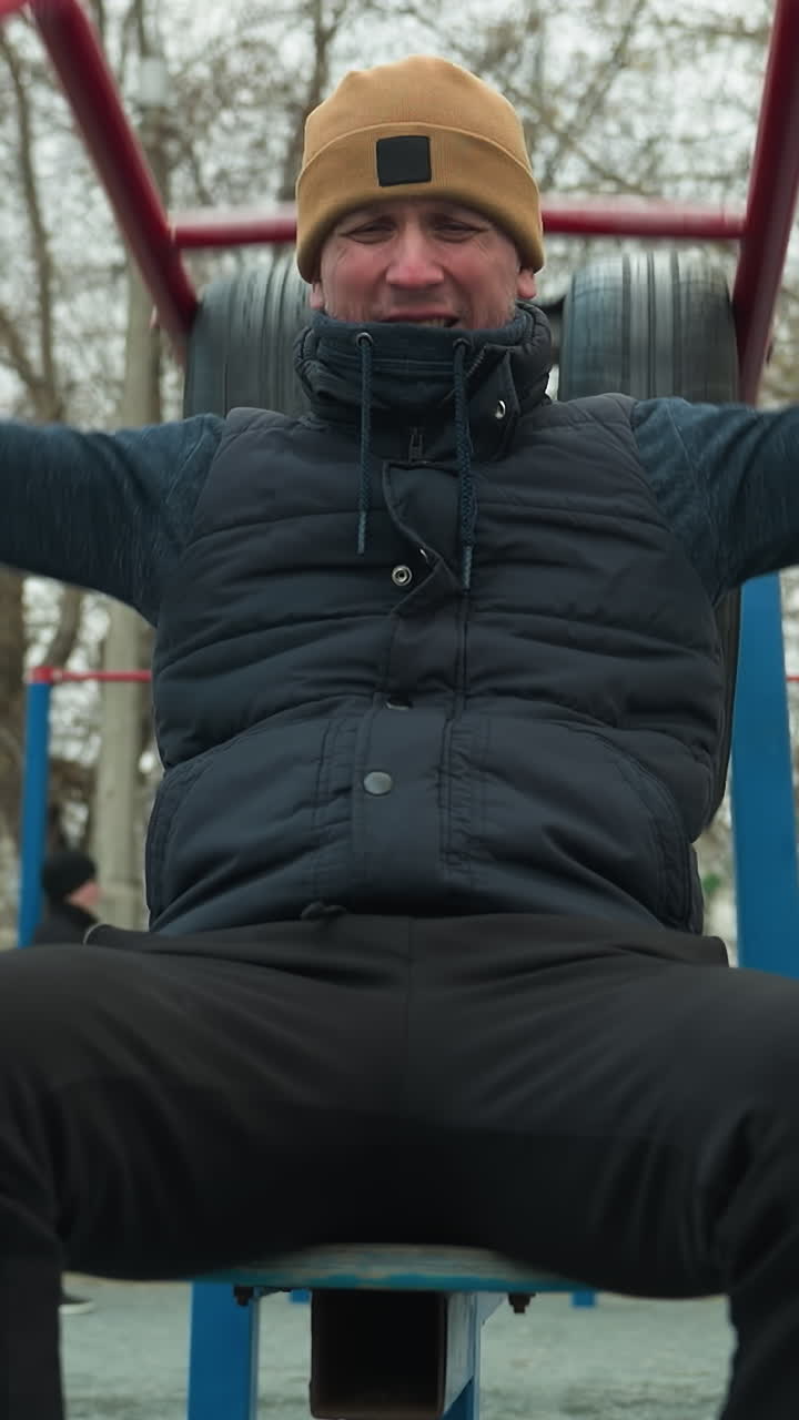 A coach wearing a brown beanie and black jacket is doing pull-ups on outdoor equipment, gripping red bars, in the background, a boy is running close to the coach
