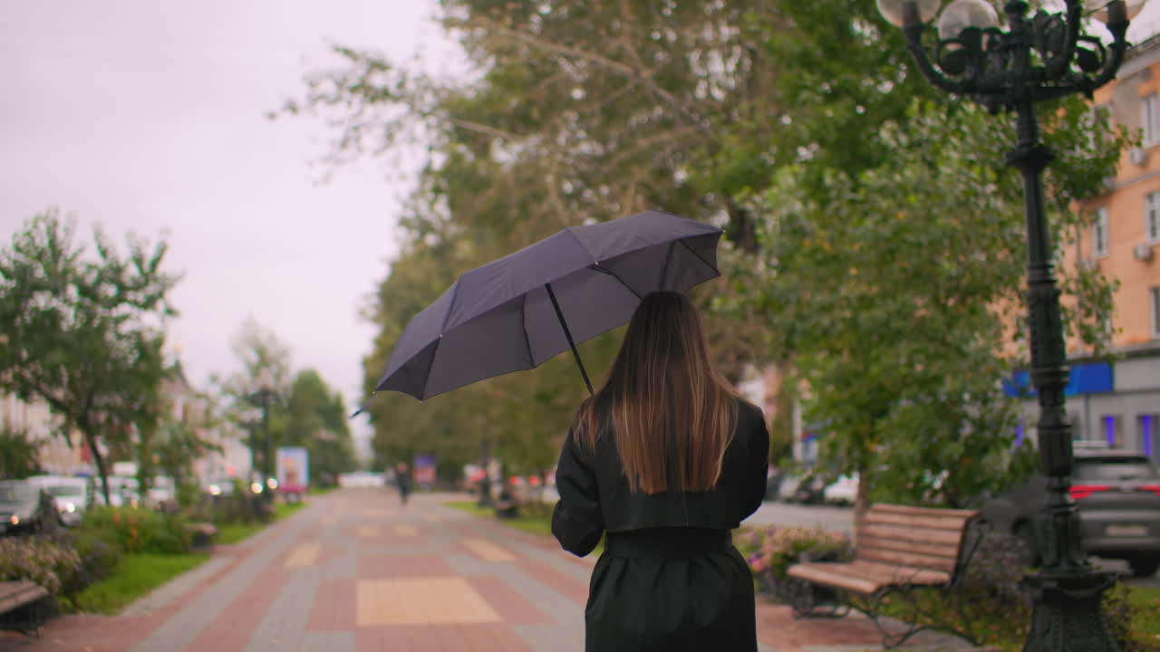 Back shot of maiden with long hair holding umbrella walking down city sidewalk on cloudy day, passing benches, trees, and street lamps, with cars in traffic lane, autumn atmosphere and urban lifestyle