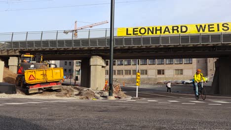 road construction at tondi bridge underpass with construction vehicles and people working during autumn day. bicycle rider passes by the camera and people are seen walking. a tall crane in the back.