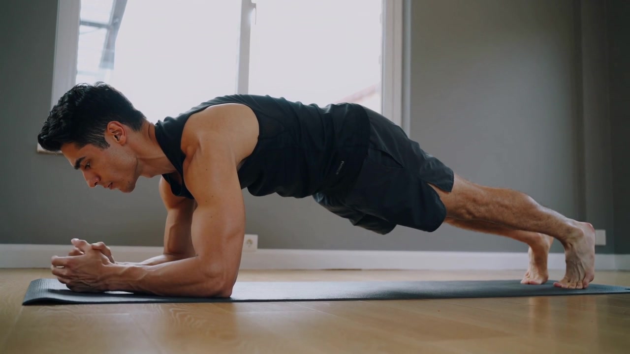 Uomo in canotta da fitness che mantiene la posizione del plank sugli avambracci dopo essersi posizionato su un tappetino nero vicino a una finestra.