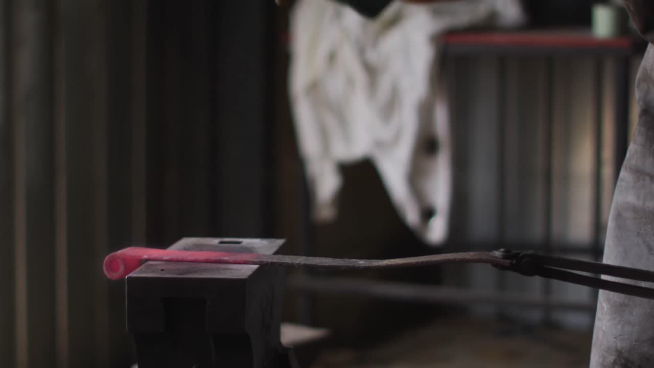 Midsection of caucasian male blacksmith wearing safety glasses, hammering hot metal tool on anvil