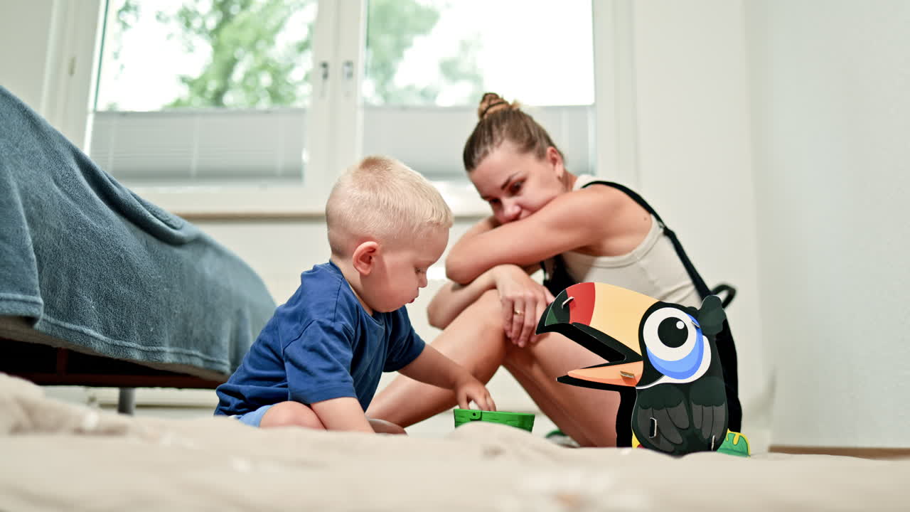 Exhausted mother sits next to a playing child. A mother in depression after giving birth sits on the floor while a child plays next to her. Postpartum depression in a woman