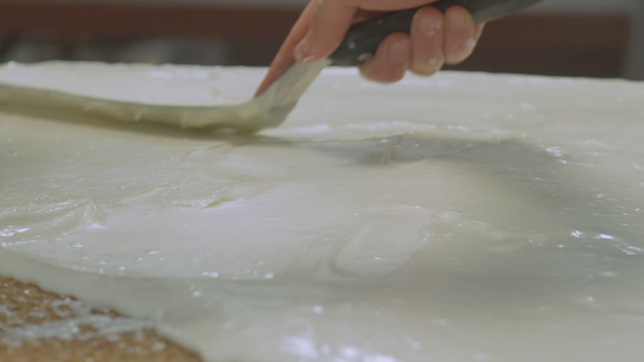 Close up of a worker spreading out icing on a large slab of cake and then decorates with a line pattern