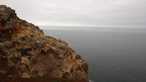 Toma-Panorámica-De-Un-Acantilado-Rocoso-Alto-Y-Dentado-Con-Un-Barco-Solitario-Navegando-En-Un-Océano-Gris-Debajo