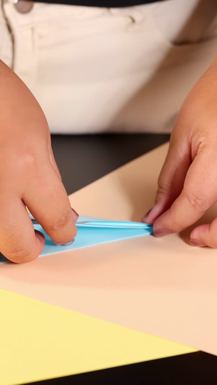 Le mani di una persona piegano con cura carta origami blu su una superficie pastello sotto un'illuminazione brillante e uniforme.