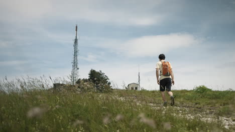 Hiker-walkih-uphill-towards-the-radio-tower-on-mountain-Slavnik,-light-wind-is-moving-grass-blades,-Clouds-on-the-blue-summer-sky