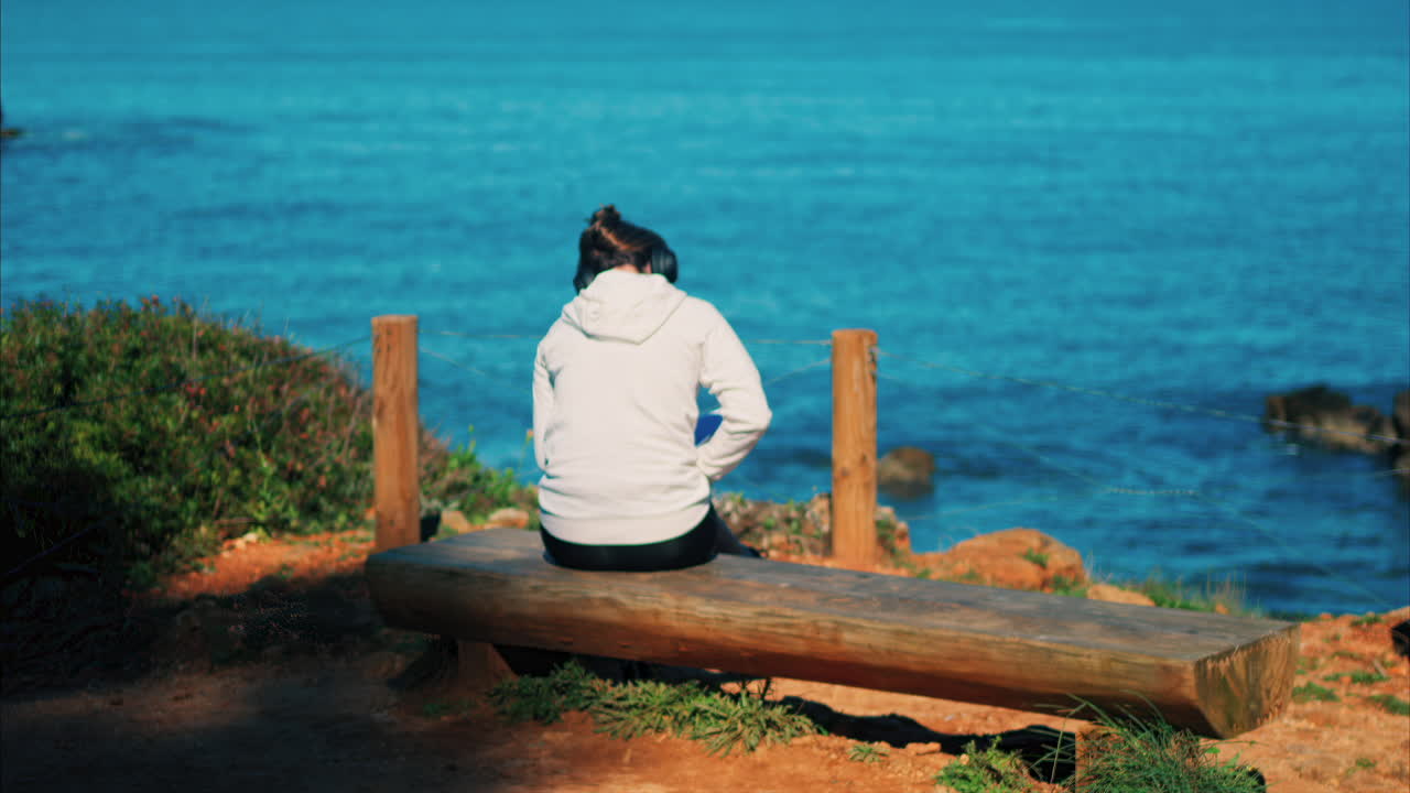 Woman with headphones on her head sitting on a wooden bench with a view of the sea