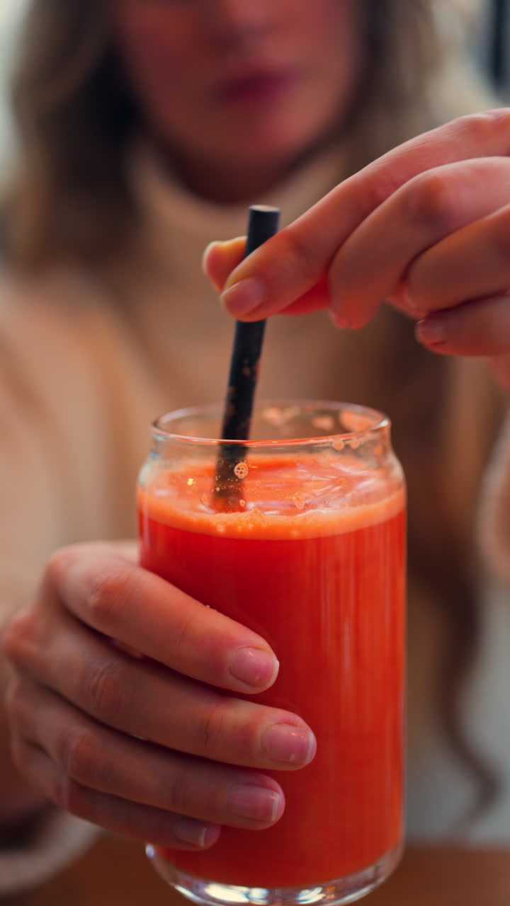 Primo piano di una donna che mescola un succo di carota e arancia in un bicchiere con una cannuccia nera su un tavolo di legno. Verticale